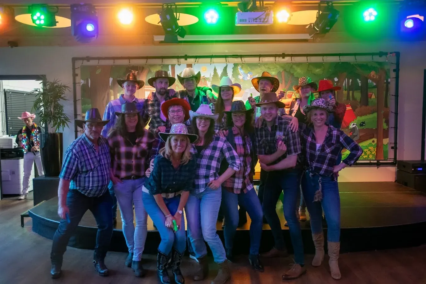 Een grote groep verkleed als cowboys en cowgirls poseert voor het podium tijdens de feestavond, met kleurrijke lichten en een beschilderd bosdecor op de achtergrond.