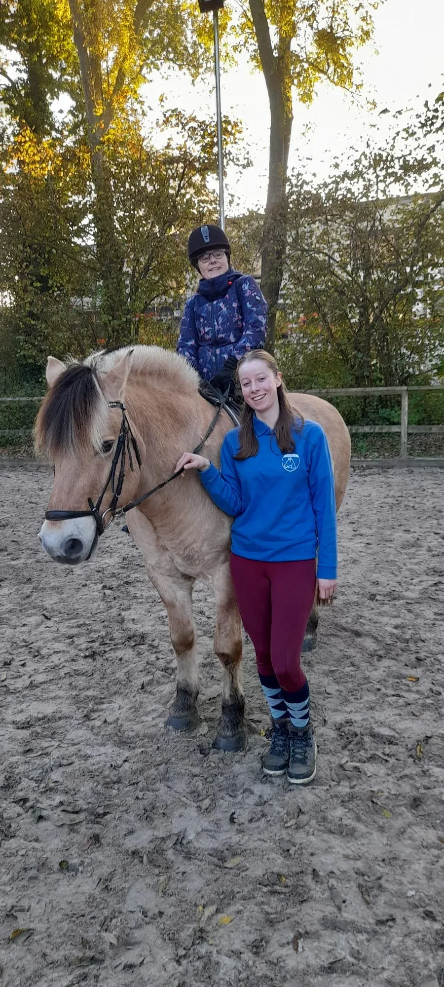 Een ruiter zit op een fjordenpaard in de buitenbak, met een vrijwilliger lachend naast het paard.