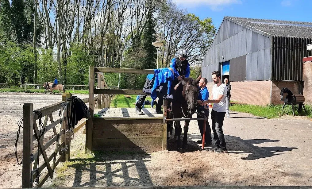 Een ruiter stapt vanaf het opstapbordes op een paard, geholpen door twee vrijwilligers.