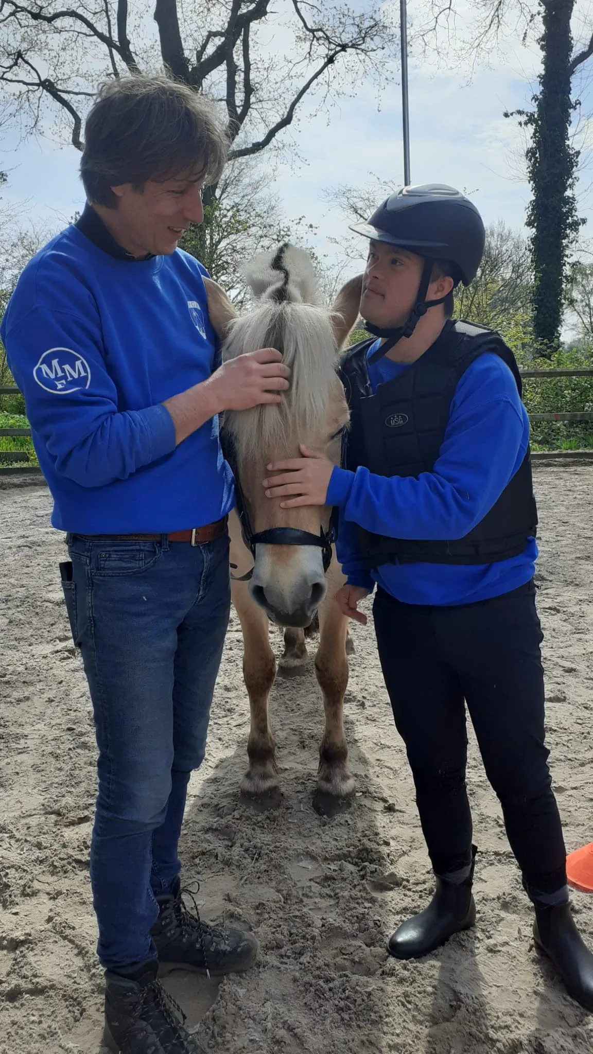 Een ruiter en een vrijwilliger aaien samen een fjordenpaard in de buitenbak.
