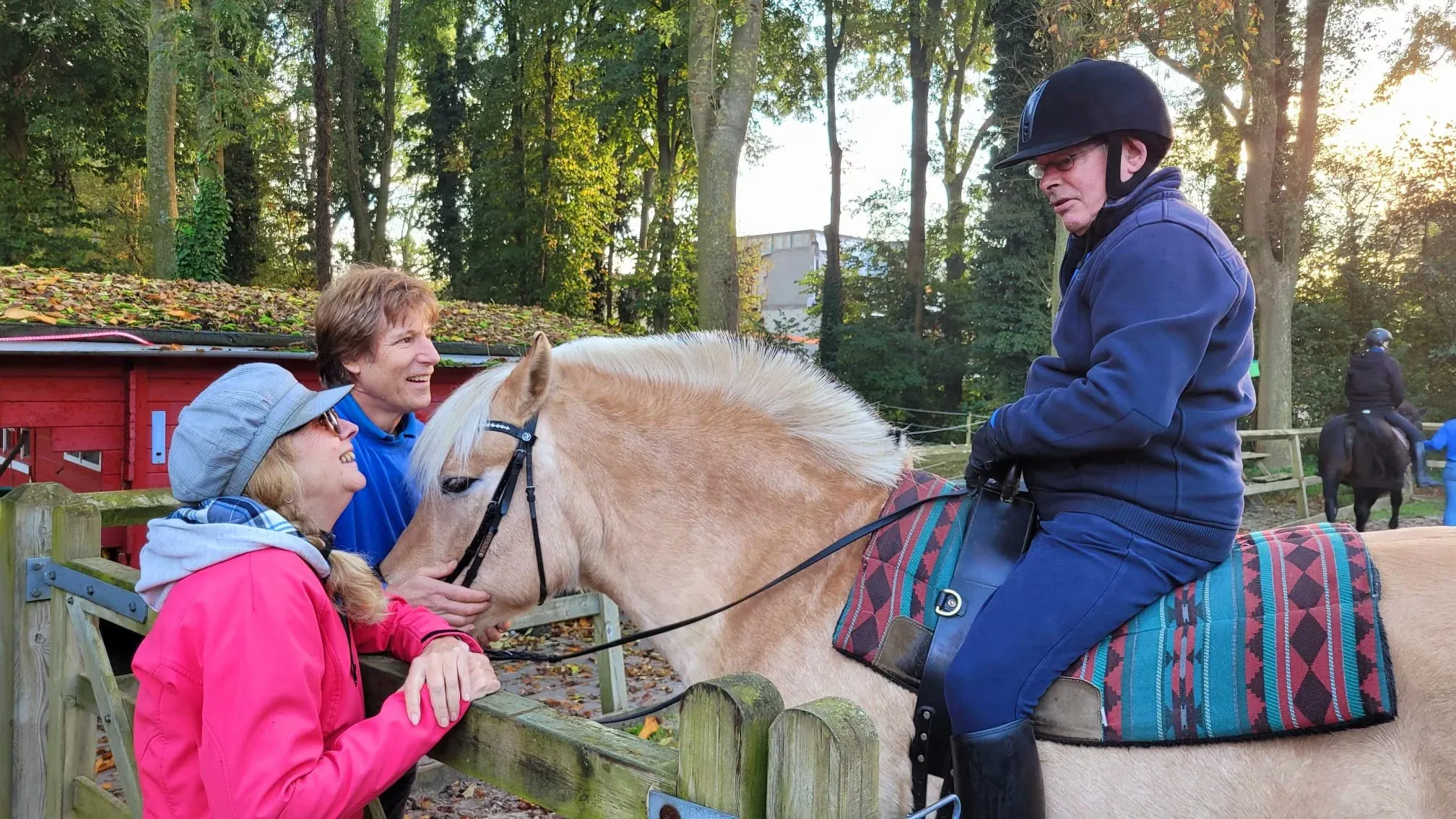 Twee vrijwilligers staan lachend bij een paard met ruiter, buiten bij de manege.